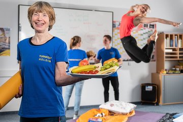Eine ältere Dame hält einen Teller mit Obst und Käse in der linken Hand. Unter dem rechten Arm trägt sie eine orange Yogamatte. Im Hintergrund machen Sportler Turnübungen.