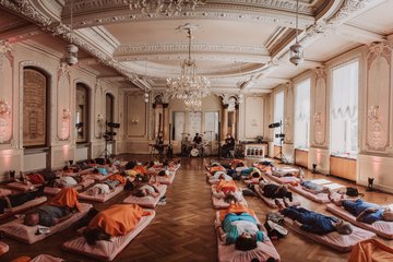 Eine Gruppe von Leuten liegt auf Matratzen in einem Barocksaal. Währendessen spielt eine Band ihre sanften Klänge.