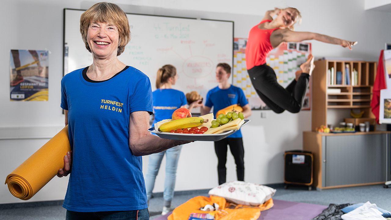 Eine ältere Dame hält einen Teller mit Obst und Käse in der linken Hand. Unter dem rechten Arm trägt sie eine orange Yogamatte. Im Hintergrund machen Sportler Turnübungen.